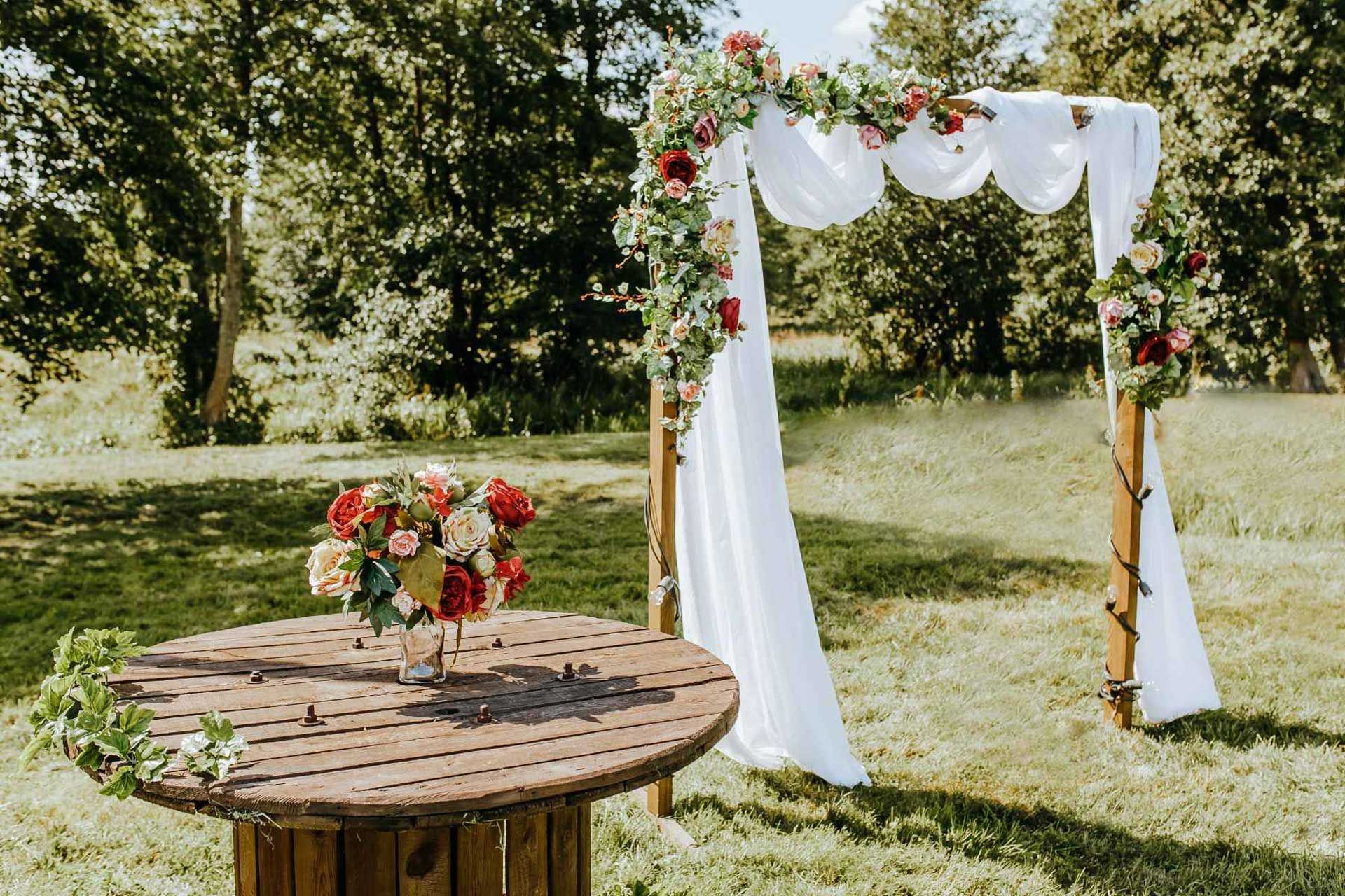 Fotografía de un prado con un arco de boda con flores, al lado de una mesa de madera con un jarrón con flores